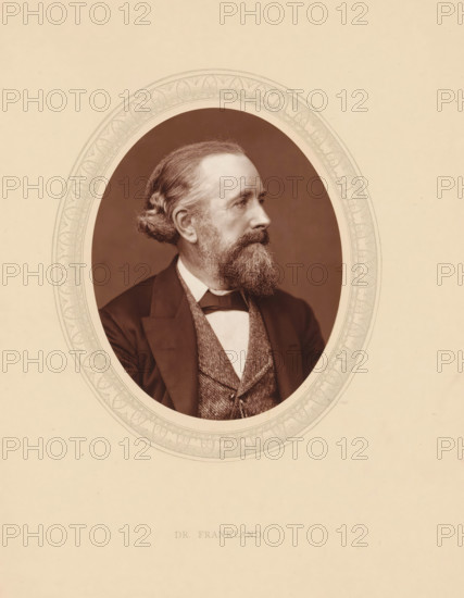 Dr. Frankland, page, photomechanical print, photographer: Lock & Whitfield, cliché maker: anonymous, c. 1875 - in or before 1880, paper, Woodburytype, Woodbury Process, height 115 mm x width 89 mm, Person, Edward Frankland, historical persons (portraits and scenes from the life) ((full) bust portrait), chemistry (portrait of scholar, scientist), A sepia-toned oval portrait of a middle-aged man shown in profile facing right, with wavy hair pulled back at the nape, a full beard and mustache, and a contemplative expression; he wears a dark jacket over a patterned waistcoat, a white shirt, and a bow tie; the portrait is set within a decorative oval border on a light beige background.