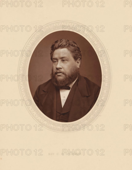Rev. CH Spurgeon, page, photomechanical print, photographer: Lock & Whitfield, cliché maker: anonymous, c. 1875 - in or before 1880, paper, Woodburytype, Woodbury Process, height 115 mm x width 89 mm, Person, Charles Haddon Spurgeon, theologian, historical persons (portraits and scenes from the life) ((full) bust portrait), An oval sepia photograph of a man with wavy hair and a full beard set within a decorative oval border on a light beige background, the man wearing a dark jacket, waistcoat, and bow tie, looking slightly to his left with a neutral expression.