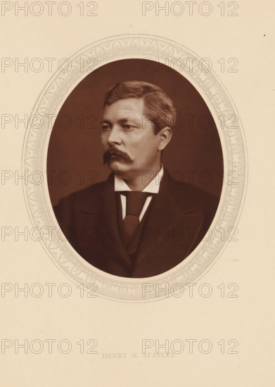 Henry M. Stanley, page, photomechanical print, photographer: Lock & Whitfield, cliché maker: anonymous, c. 1875 - in or before 1880, paper, Woodburytype, Woodbury Process, height 115 mm x width 89 mm, Person, sir Henry Morton Stanley, explorer, historical persons (portraits and scenes from the life) ((full) bust portrait), An oval sepia portrait of a man with neatly combed hair and a prominent mustache set against a dark background, framed by an ornate patterned border on a light beige sheet, the man wearing a dark suit jacket, waistcoat, high starched collar, and a tied cravat, his face turned slightly to the viewer’s left with a neutral expression.