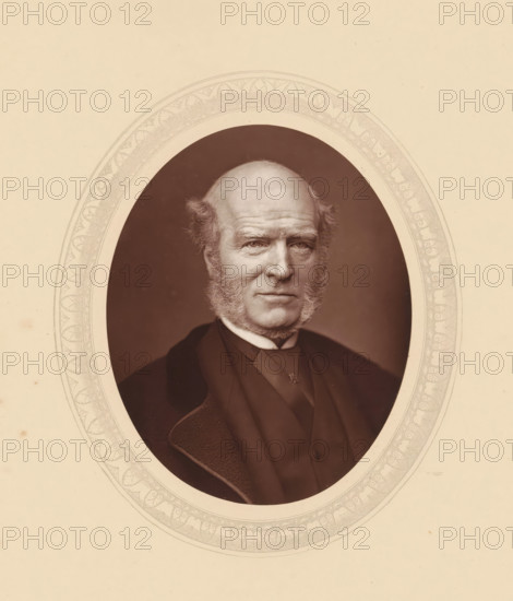 Thomas Hughes, page, photomechanical print, photographer: Lock & Whitfield, cliché maker: anonymous, c. 1875 - in or before 1880, paper, Woodburytype, Woodbury Process, height 115 mm x width 89 mm, Person, lawyer, attorney at law, historical persons (portraits and scenes from the life) ((full) bust portrait), An oval sepia photograph of an older man with a high forehead and receding hairline, prominent sideburns, and a serious expression, shown from the chest up wearing a dark coat, waistcoat, and a cravat with a small ornamental pin, set against a dark background and surrounded by a decorative patterned oval mat on a pale paper.