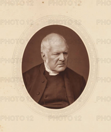 Edward Meyrick Goulburn, DD, page, photomechanical print, photographer: Lock & Whitfield, cliché maker: anonymous, c. 1875 - in or before 1880, paper, Woodburytype, Woodbury Process, height 115 mm x width 89 mm, Person, theologian, historical persons (portraits and scenes from the life) ((full) bust portrait), An oval sepia portrait of an older man with light hair and sideburns set against a dark background, showing his face in three-quarter view with a serious expression, dressed in a dark coat and a high white collar, framed by an ornate pale oval mat with decorative patterns and surrounded by a light, slightly spotted background.