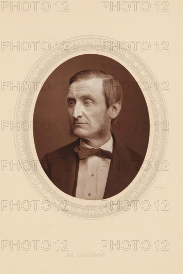 Dr. Gladstone, page, photomechanical print, photographer: Lock & Whitfield, cliché maker: anonymous, c. 1875 - in or before 1880, paper, Woodburytype, Woodbury Process, height 115 mm x width 89 mm, Person, John Hall Gladstone, historical persons (portraits and scenes from the life) ((full) bust portrait), chemistry (portrait of scholar, scientist), An oval portrait of a man with sideburns and short, combed hair, shown from the chest up and turned slightly to his left, wearing a dark jacket, a light shirt with a high collar, and a bow tie, set against a dark background and framed by an ornate decorative oval mat on a light paper background.