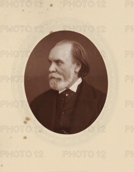 Sir Theodore Martin, page, photomechanical print, photographer: Lock & Whitfield, cliché maker: anonymous, c. 1876 - in or before 1881, paper, Woodburytype, Woodbury Process, height 115 mm x width 89 mm, Person, Theodore Martin, portrait of a writer, historical persons (portraits and scenes from the life) ((full) bust portrait), A sepia-toned oval portrait of an older man shown in three-quarter profile facing left, with a full beard and mustache, a high forehead with receding hairline, and hair swept back; he wears a dark suit jacket over a vest and a patterned cravat with a white collar peeking out, and the portrait is set against a dark background within a decorative oval mat that shows faint embossed patterns and a few small stains on the surrounding light-colored paper.