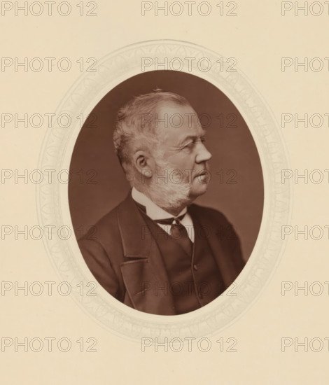 Duke of Richmond and Gordon, page, photomechanical print, photographer: Lock & Whitfield, cliché maker: anonymous, c. 1877 - in or before 1882, paper, Woodburytype, Woodbury Process, height 115 mm x width 89 mm, Person, Charles Gordon-Lennox, politician, eg party leader, historical persons (portraits and scenes from the life) ((full) bust portrait), A sepia-toned oval portrait of an older man shown in profile facing right, with short swept-back hair and prominent sideburns, wearing a dark suit jacket, waistcoat, collared shirt, and a tied neckwear; the portrait is centered within a decorative oval mat with a patterned border and set against a plain light background.