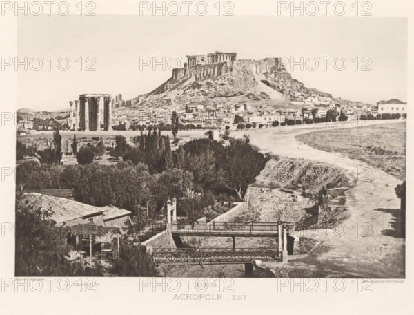 View of the Acropolis of Athens, Acropole-est, page, photomechanical print, Athens, cliché maker: Dujardin, printer (person): Chardon-Wittmann, c. 1880 - in or before 1890, photographer, printer (person), Periféreia Protevoúsis, paper, photoaquatint, height 145 mm x width 215 mm, city-view in general, 'veduta', ruin of a building, architecture, Akropolis, A wide sepia-toned landscape showing a rocky hill topped by ancient columned ruins overlooking a town with clustered low buildings and scattered trees, a group of tall standing columns visible on the plain near the hill, a winding dry riverbed running through the scene, foreground vegetation and tiled rooftops, and a pair of iron footbridges crossing a sunken channel.