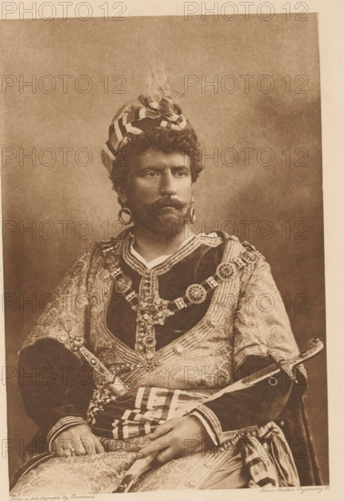 Wilson Barrett (Othello), page, photomechanical print, photographer: Herbert Rose Barraud, cliché maker: Swan Electric Engraving Co., c. 1880 - in or before 1890, paper, photoaquatint, height 191 mm x width 127 mm, Person, portrait of actor, actress, A sepia-toned portrait of a seated man wearing a striped turban with a feather, large circular earrings, a curled mustache and beard, and a richly patterned robe adorned with ornate chains and medallions, his hands resting in his lap over a patterned cloth and a sheathed sword visible at his side.