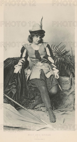Miss Ada Rehan (Rosalind), page, photomechanical print, photographer: Alexander Bassano, cliché maker: anonymous, c. 1880 - in or before 1890, paper, photoaquatint, height 203 mm x width 135 mm, Person, Ada Rehan, portrait of actor, actress, A seated person wearing a feathered hat and a costume with laced bodice and puffed sleeves appears in a monochrome photograph, resting one arm on a draped dark fabric and the other on a stone ledge near a cluster of long, narrow-leaved plants; the outfit includes a wide belt with attached pouches, a short skirt over fitted stockings, and laced ankle boots, and a long staff or pole lies diagonally across the foreground while fallen leaves and textured fabric cover the ground.
