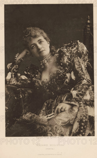 Madame Modjeska (Portia), page, photomechanical print, photographer: Benjamin Joseph Falk, New York (city), cliché maker: anonymous, publisher: Virtue & Co., London, c. 1880 - in or before 1890, North America, paper, photoaquatint, height 206 mm x width 137 mm, Person, Helena Modjeska, portrait of actor, actress, A seated woman wearing an ornate, patterned dress with puffed, decorated sleeves and lace details leans her head on one hand and looks slightly upward; she has curled hair adorned with a small hair ornament and wears a multi-strand necklace with dangling elements; her other hand rests on her lap holding a folded fan, and a feathered accessory lies nearby; the background is dark and plain and a carved wooden chair back is visible behind her.