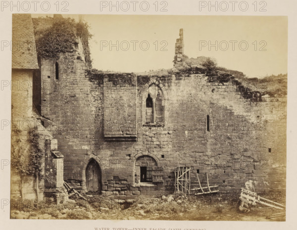Inner wall of the ruin of a water tower of Kenilworth Castle, Water Tower - Inner façade (13th Century), photograph, page, Kenilworth Castle, c. 1862 - in or before 1872, photographer, Kenilworth, paper, albumen print, height 152 mm x width 204 mm, ruin of a dwelling, house, castle, water tower, A weathered stone wall with irregular rectangular blocks, showing areas of darker discoloration and patches of vegetation growing on and over the upper edge, contains a narrow arched window high up and a larger pointed arched opening below with a wooden door set into it, a square barred window with a wooden shutter sits beside the lower arch, and leaning wooden ladders and racks rest against the wall near the ground amid scattered stones and rubble.