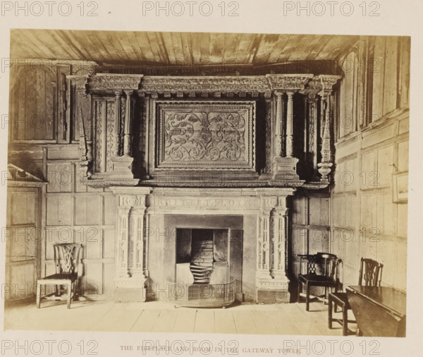 Fireplace in Kenilworth Castle, The Fireplace and Room in the Gateway Tower, photograph, page, Kenilworth Castle, c. 1862 - in or before 1872, photographer, Kenilworth, paper, albumen print, height 152 mm x width 202 mm, open hearth, fire-place, ruin of a dwelling, house, castle, A large carved stone fireplace with an ornate overmantel featuring floral and scrollwork panels is centered in a paneled room, flanked by decorative pilasters and carved columns supporting a heavily carved frieze. The fireplace opening shows stacked bricks and a metal fireguard curved around the hearth. Wooden wall paneling covers the surrounding walls and a plank ceiling runs overhead. Simple wooden chairs with carved backs and a small wooden table are placed near the fireplace on a wide wooden plank floor.