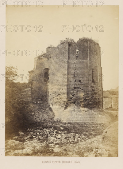 Ruin of Lunn's Tower of Kenilworth Castle, Lunn's Tower (before 1200), photograph, page, Kenilworth Castle, c. 1862 - in or before 1872, photographer, Kenilworth, paper, albumen print, height 204 mm x width 152 mm, ruin of a dwelling, house, castle, tower, clock tower, monumental adornment, A tall circular stone tower with a battered base and visible horizontal courses shows areas of missing and crumbling masonry and a narrow vertical slit opening with vegetation poking through; adjacent to it is a partially ruined wall with an arched gap and jagged top, and rubble and scattered stones lie on grassy earth in the foreground while low vegetation and a tree appear to the left.
