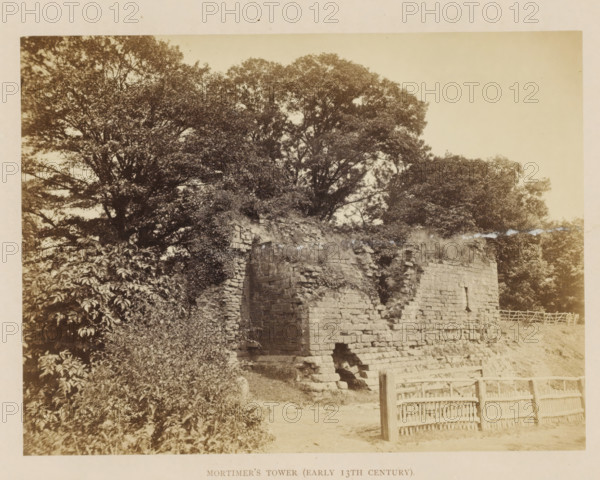 View of the ruins of Mortimer's Tower at Kenilworth Castle, Mortimer's Tower (Early 13th Century), photograph, page, Kenilworth Castle, c. 1862 - in or before 1872, photographer, Kenilworth, paper, albumen print, height 152 mm x width 204 mm, ruin of a dwelling, house, castle, tower, clock tower, monumental adornment, A sepia-toned photograph shows a partially ruined stone wall with large rough blocks and an opening near its base, vegetation growing on and around the masonry, dense trees with broad canopies rising behind the wall, shrubs and climbing plants covering parts of the left side, a simple wooden fence with vertical and horizontal slats in the foreground, and a dirt path leading toward the wall.