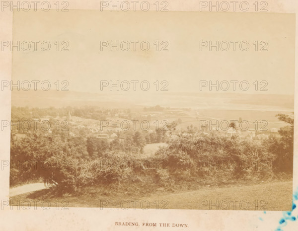 Brading, from the Down, photograph, page, photographer: William Russell Sedgfield, Isle of Wight, c. 1859 - in or before 1869, paper, albumen print, height 100 mm x width 144 mm, Photo on frontispiece., city-view in general, 'veduta', A faded sepia-toned panoramic view showing a landscape with a foreground of grassy slopes and dense scrubby bushes, a midground with clusters of trees and scattered buildings including a structure with a steeple, and a distant horizon of low rolling hills and open fields under a pale sky.