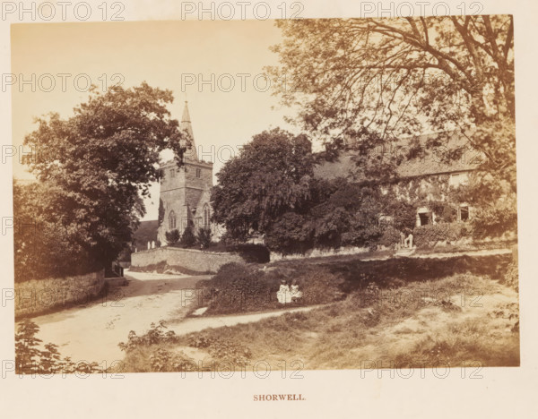 Shorwell, photograph, page, photographer: William Russell Sedgfield, c. 1859 - in or before 1869, Isle of Wight, paper, albumen print, height 100 mm x width 143 mm, public gardens, park, church (exterior), A sepia-toned outdoor scene showing a stone church with a square tower and a tall pointed spire partially obscured by large leafy trees, a stone wall curving along a dirt path in front of the church, a sloped grassy area and low shrubs in the foreground, a large tree with spreading branches on the right casting dappled shade over a building with multiple windows partially covered by climbing plants, stone steps leading up to the building where two standing figures are visible near the entrance, and a small group of seated figures or statues on the grassy bank near the path.