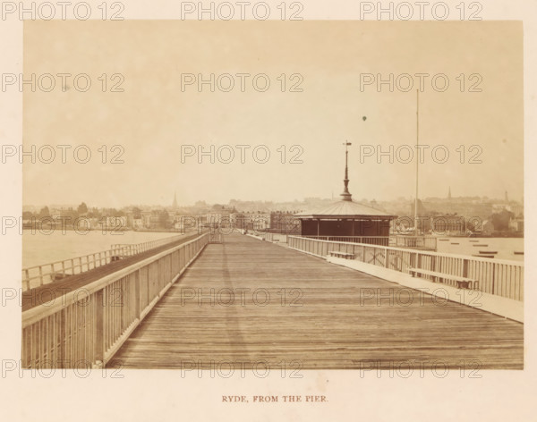 Ryde, from the pier, photograph, page, photographer: William Russell Sedgfield, c. 1859 - in or before 1869, Isle of Wight, paper, albumen print, height 101 mm x width 144 mm, city-view in general, 'veduta', pier, quay, wharf, A wide wooden pier extends toward a distant shoreline, with wooden planks forming the walking surface and railings running along both sides; a pavilion with a pointed central finial and a column rises on the right side of the pier, and a tall thin mast stands beyond it; the shoreline shows rows of buildings and trees with various rooflines and a few church spires faintly visible against a pale sky; calm water borders the left side of the pier and small boats appear near the right shoreline; the overall image has a sepia tone and an aged, slightly faded appearance.