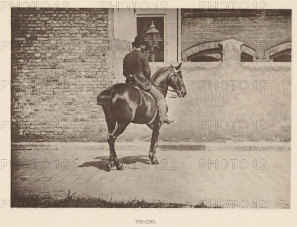 Man on a horse in a travers, Travers, page, photomechanical print, photographer: Alexander Nicol (possibly), Edinburgh (possibly), cliché maker: Römmler & Jonas, Dresden, c. 1879 - in or before 1889, paper, collotype, photoprint, height 98 mm x width 138 mm, horse, riding a horse, ass, or mule, rider, horseman, head-gear: bowler hat, A person wearing a dark coat and a hat sits astride a dark horse, viewed from behind and slightly to the side, the horse showing a glossy coat and a visible bridle; they are positioned on a paved surface in front of a wall that has a rough lower portion and a brick upper portion with an arched brick feature and a window partially visible above the rider, and a lamp post stands near the window.