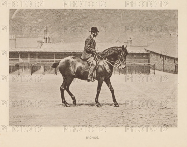 Backing, Man on a horse walking backwards, page, photomechanical print, photographer: Alexander Nicol (possibly), Edinburgh (possibly), cliché maker: Römmler & Jonas, Dresden, c. 1879 - in or before 1889, paper, collotype, photoprint, height 96 mm x width 137 mm, horse, riding a horse, ass, or mule, rider, horseman, head-gear: bowler hat, A sepia-toned photograph of a person seated on a horse in an open sandy area, the rider wearing a dark bowler hat and a jacket while holding the reins, the horse shown in profile with a bridle and a sturdy build, a low fence and a long building visible in the background under a textured sky.