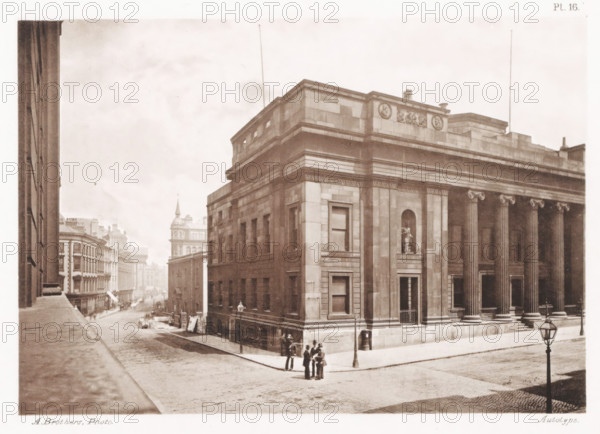 View of the public library in Manchester, located in the old town hall, The free public Library, Old Town Hall, page, photomechanical print, number: 'Pl. 16.', photographer: Brothers, Alfred, Manchester, cliché maker: anonymous, c. 1868 - in or before 1878, printer (person), paper, collotype, photoprint, height 137 mm x width 197 mm, public library, A sepia-toned street scene shows a large stone building with classical columns along its facade and a recessed niche containing a statue, with ornamental friezes and a flat roofline; a group of people stands on the corner near a lamppost while other figures are scattered along the pavement; the cobbled street stretches away past rows of multistory buildings with varied rooflines and a distant tower with a pointed roof; a tall lamppost is visible in the foreground and another near the building, with architectural details such as windows, cornices, and stone blocks evident on the main structure.