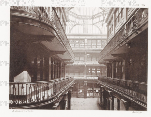 View of the Barton Arcade in Manchester, Barton Arcade, page, photomechanical print, number: 'Pl. 29.', photographer: Brothers, Alfred, Manchester, cliché maker: anonymous, c. 1868 - in or before 1878, printer (person), paper, collotype, photoprint, height 142 mm x width 190 mm, shop, store, arcade, architecture, A sepia-toned interior view showing a tall atrium with ornate iron balustrades curving along upper walkways on both sides and decorative moldings along the walls, a large glass skylight with metal framing forming geometric arches at the top left, rows of windows and doorways along the back wall, display cases and shelving visible on the ground level, patterned shadows cast across floors and walls, and overall symmetrical architectural details extending vertically through the space.