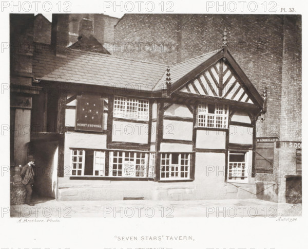 Seven Stars' Tavern, Shudehill, View of an inn in Manchester, page, photomechanical print, number: 'Pl. 33.', photographer: Brothers, Alfred, Manchester, cliché maker: anonymous, printer (person): Brothers, c. 1868 - in or before 1878, Brothers, photographer, cliché maker, printer (person), paper, collotype, photoprint, height 133 mm x width 191 mm, Print after page 30., hotel, hostelry, inn, A two-story timber-framed building with dark wooden beams forming a pattern against lighter plaster panels, a steep tiled roof with decorative finials at the gable peak, multiple leaded-paned windows across both stories including a projecting bay window, an arched entrance on the left where a man in a suit and hat leans against the wall, a brick wall rising behind the building, and a paved area in front.