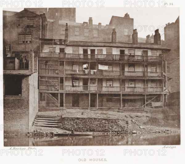 View of a house in Manchester with an open gallery, Old houses, parsonage, page, photomechanical print, number: 'Pl. 34.', photographer: Brothers, Alfred, Manchester, cliché maker: anonymous, c. 1868 - in or before 1878, printer (person), paper, collotype, photoprint, height 140 mm x width 179 mm, Print after page 30., housing, A multi-story brick building with wooden balconies running across several levels, each balcony supported by vertical posts and accessed by an exterior wooden staircase, clothes hanging over one railing, multiple small paned windows set into the facade, chimney stacks rising from the roofline, adjoining taller brick buildings visible behind and beside it, a stone terraced embankment leading down to a calm body of water in the foreground with a small boat pulled up at the edge, exposed earth and scattered debris along the shoreline.