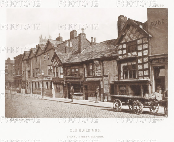 View of Chapel Street in Salford, Old buildings, Chapel Street, Salford, page, photomechanical print, number: 'Pl. 39.', photographer: Brothers, Alfred, cliché maker: anonymous, c. 1868 - in or before 1878, printer (person), paper, collotype, photoprint, height 125 mm x width 187 mm, street, four-wheeled, animal-drawn vehicle, eg: cab, carriage, coach, A sepia-toned street scene showing a row of old timber-framed and brick buildings with steep gabled roofs and multiple chimneys, windows of various sizes and pane arrangements, and exposed wooden beams and plaster panels on the facades; two men stand on the sidewalk near building entrances, one facing a shop window and the other standing by a doorway beneath posters on a wall; a wooden cart with large spoked wheels is parked at the curb in front of the buildings; a cobblestone street runs along the front and a single lamp post stands near the pavement edge; a distant church tower with pointed spires is visible above the rooftops in the b...