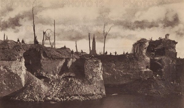 View of a breach in a bastion in Strasbourg as a result of the siege by the German army, Face gauche de la brèche du bastion 12 et porte de pierres, page, photomechanical print, photographer: Charles David Winter, Strasbourg (France), cliché maker: anonymous, printer (person): Goupil & Cie., 1870, printer (person), paper, Woodburytype, height 101 mm x width 170 mm, Print on frontispiece., landscape with ruins, assault, siege, consequences of war, A wide, sepia-toned scene shows a ruined landscape with crumbling stone walls and mounds of rubble descending to a dark body of water in the foreground, jagged exposed brick and arch shapes visible in the ruins. On the higher ground above the ruins are silhouettes of bare, leafless tree trunks and sparse branches rising against a cloudy sky. Small human figures stand and sit along the ridge, their forms dark and indistinct. On the right a partially collapsed building with broken beams and torn masonry leans over the edge of the ruin, with scat...