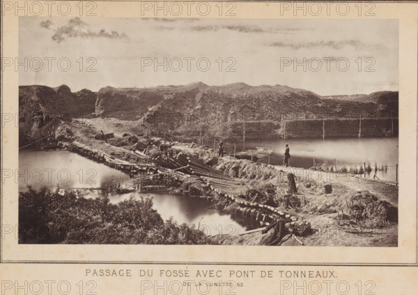 Passage du fossé avec pont de tonneaux de la lunette 52, View of a passage through a fortress and a pontoon bridge in Strasbourg, page, photomechanical print, photographer: Charles David Winter, Strasbourg (France), cliché maker: anonymous, printer (person): Goupil & Cie., 1870, printer (person), paper, Woodburytype, Woodburytypie, height 101 mm x width 171 mm, assault, siege, pontoon bridge, consequences of war, A sepia-toned photograph showing a rough wooden bridge and pathway crossing a body of water with reflections, flanked by earthworks and uneven terrain; the foreground includes dense foliage and scattered wooden debris, while the middle ground features men standing on the path near small boats tied at the water’s edge and ropes or posts lining the route; the background shows rocky, grassy embankments rising to a ridge under a sky with light cloud streaks.
