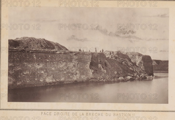 Face droite de la brèche du bastion 11, Gezicht op een bres in een bastion te Straatsburg tengevolge van een besiege door het Duitse leger, page, photomechanical print, photographer: Charles David Winter, Straatsburg (France), clichémaker: anonymous, printer (person): Goupil & Cie., 1870, printer (person), paper, Woodburytype, Woodburytypie, height 101 mm x width 171 mm, bastion, bulwark, assault, siege, consequences of war, A wide sepia-toned photograph shows a large stone and earth embankment rising from a calm body of water, with the embankment surface textured by horizontal lines and patches of lighter discoloration. The top edge of the embankment is uneven and rocky, and a group of small human figures stand and sit along the ridge and slopes, some near areas of exposed rubble. To the left a rounded rocky hill rises behind the embankment, and to the right the embankment steps down into a steep, broken cliff face with scattered rocks at its base. The sky above is pale with faint, wi...
