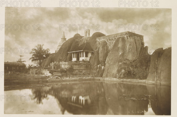 Monolithic cistern at Anuradhapura, Sri Lanka, page, photomechanical print, signature: 'Woodbury P ?', title: 'MONOLITHIC CISTERN.', photographer: Henry William Cave, Anuradhapura, cliché maker: Woodbury Permanent Photographic Printing Company, 1896, South Asia, paper, autotypie, height 76 mm x width 120 mm, sink, basin, Thuparamaya, A sepia-toned scene shows a stone structure set against large rounded rock formations beside a calm reflective pool, with a small pavilion featuring a gabled roof and open sides perched on a raised stone platform; there are carved stone steps and remnants of masonry leading down to the water, a tall palm tree and other vegetation near the rocks, and a stepped stone terrace and wall extending along the right side, while distant temple-like spires and an archway rise behind the rocks beneath a cloudy sky.