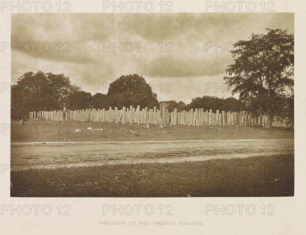 Entrance to the Bodhi tree enclosure, Anuradhapura, Sri Lanka, page, photomechanical print, title: 'ENTRANCE TO THE BO-TREE ENCLOSURE.', photographer: Henry William Cave, cliché maker: anonymous, 1896, South Asia, paper, Woodburygravure, height 97 mm x width 146 mm, Print for page 45., sculpture, places and objects of worship, Hinduism, Buddhism, Jainism, trees: fig-tree, A sepia-toned photograph showing a grassy foreground with a dirt track or worn strip running across it, a long irregular line of upright wooden posts of varying heights behind the strip, scattered pieces of wood and stones on the ground near the posts, clusters of large leafy trees rising behind and to the sides of the posts, a cloudy sky filling the upper portion of the image, and a narrow pale border around the photograph.