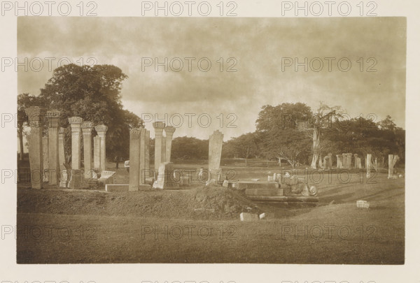 Ruins near the Ruanwelisaya stupa at Anuradhapura, Sri Lanka, page, photomechanical print, title: 'RUINS NEAR THE RUANWELI DAGABA.', photographer: Henry William Cave, Anuradhapura, cliché maker: anonymous, 1896, South Asia, paper, Woodburygravure, height 124 mm x width 195 mm, Print for page 65., landscape with ruins, column, pillar, architecture, places and objects of worship, Hinduism, Buddhism, Jainism, A sepia-toned landscape showing a grassy foreground and a cluster of tall stone columns and carved architectural fragments arranged on uneven ground, some columns standing upright and others leaning or broken, with decorative capitals and sculpted bases visible; a coiled ornamental stone piece lies near the front among scattered blocks and carved slabs; behind the ruins a line of trees with dense foliage stretches across the middle distance, including one prominent bare-trunked tree, and a wide cloudy sky fills the upper portion of the scene.
