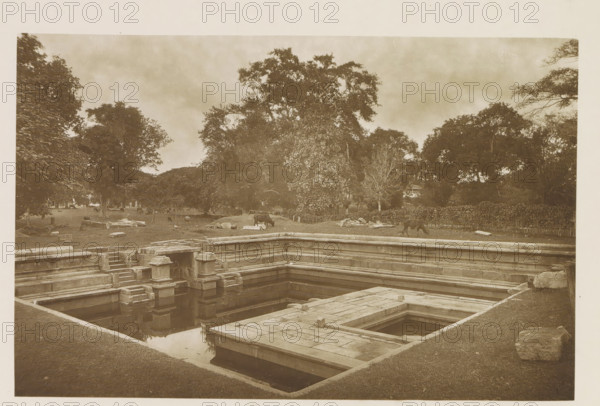Pond near the Ruwanwelisaya stupa in Anuradhapura, Sri Lanka, page, photomechanical print, title: 'POKUNA.', photographer: Henry William Cave, Anuradhapura, clichémaker: anonymous, 1896, South Asia, paper, Woodburygravure, height 125 mm x width 193 mm, Print for page 67., pond, pool, places and objects of worship, Hinduism, Buddhism, Jainism, A stone stepped rectangular pool with layered ledges and square platforms sits in the foreground, its water reflecting the stonework; beyond the pool a grassy area with scattered stones and low piles leads to a line of trees with dense foliage, and a few grazing animals are visible near the treeline; tall trees with broad canopies fill the middle and background under a cloudy sky.