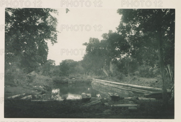 View of the Kuttam Pokuna ponds near the Ruwanwelisaya stupa in Anuradhapura, Sri Lanka, page, photomechanical print, title: 'KUTTAM-POKUNA.', photographer: Henry William Cave, Anuradhapura, cliché maker: anonymous, 1896, South Asia, paper, Woodburygravure, height 126 mm x width 194 mm, Print for page 69., pond, pool, places and objects of worship, Hinduism, Buddhism, Jainism, A black-and-white photograph of a wooded area showing dense tree canopies framing an open sky, a calm rectangular pool of water reflecting surrounding foliage, a low stone wall running along one side of the pool, scattered stone blocks and circular stone features near the water's edge, overgrown vegetation and shrubs around the pool, and tree trunks and branches visible in the foreground and background.
