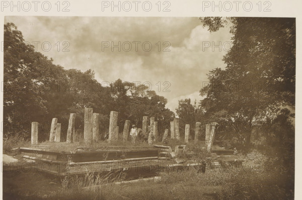 Ruins of a pavillion at Anuradhapura, Sri Lanka, page, photomechanical print, title: 'REMAINS OF PAVILLION (THIRD CENTURY).', photographer: Henry William Cave, Anuradhapura, cliché maker: anonymous, 1896, South Asia, paper, Woodburygravure, height 124 mm x width 193 mm, Print for page 79., ruin of a building, architecture, column, pillar, A sepia-toned outdoor scene shows a grassy area with a series of upright stone pillars of varying heights arranged in a loose formation, surrounded by low stone foundations and scattered rectangular stone blocks. A solitary person stands among the pillars wearing light clothing and a head covering. Dense trees frame the background on both sides, and a cloudy sky fills the upper portion of the image. The ground in the foreground has grasses and low vegetation interspersed with the stone structures.