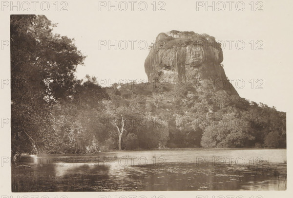 View of the Lion Rock at Sigiriya, Sri Lanka, page, photomechanical print, title: 'SIGIRI.', photographer: Henry William Cave, Sigiriya, cliché maker: anonymous, 1896, South Asia, paper, Woodburygravure, height 126 mm x width 195 mm, Print for page 93., lake, rock formations, A large rocky outcrop rises from a densely treed hillside, its steep face showing vertical striations and a rounded top with scattered trees. In front of the hill, a band of forest with varied tree shapes fills the middle ground, including a tall bare-trunked tree that stands out against the foliage. A body of water occupies the foreground, its surface reflecting light and showing small patches of aquatic vegetation. On the left edge, overhanging branches and dense foliage frame the scene. The image is monochrome and framed with a narrow light border.