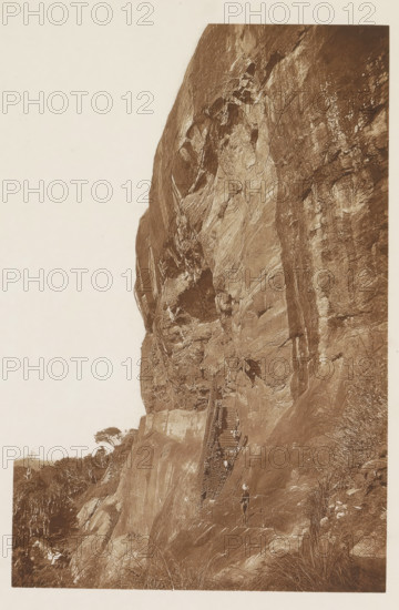 View of the gallery at the Lion Rock, Sigiriya, Sri Lanka, page, photomechanical print, title: 'REMAINS OF THE GALLERIES OF SIGIRI.', photographer: Henry William Cave, cliché maker: anonymous, 1896, South Asia, paper, Woodburygravure, height 191 mm x width 125 mm, Print for page 95., ruin of a dwelling, house, castle, rock-formations, A steep rock face fills most of the image, showing weathered stone surfaces with visible cracks, ledges, and textured striations. A narrow wooden staircase with railings ascends along the rock, anchored to the stone, and several small figures stand on and near the stairs. Vegetation including low grasses and scattered trees appears at the base and along the left edge of the scene. The photograph is sepia-toned and framed with a pale margin.