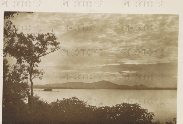 View of the water reservoir at Minneriya, Sri Lanka, page, photomechanical print, title: 'MINNERIA.', photographer: Henry William Cave, Minneriya, cliché maker: anonymous, 1896, South Asia, paper, Woodburygravure, height 126 mm x width 195 mm, Print for page 101., dammed-up lake, A sepia-toned landscape showing a calm body of water stretching toward distant hills under a cloudy sky, a tall tree with leafy branches silhouetted on the left edge, low shrubbery in the foreground, and a small island or rocky outcrop near the shoreline.