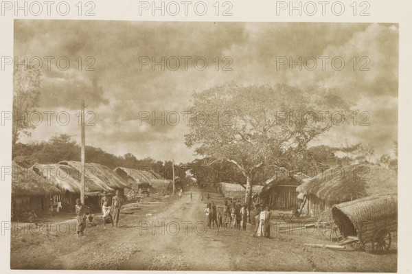 Path through Kekirawa, Sri Lanka, page, photomechanical print, title: 'KEKIRAWA.', photographer: Henry William Cave, Kekirawa, cliché maker: anonymous, 1896, South Asia, paper, Woodburygravure, height 124 mm x width 192 mm, Print for page 21., public road in village, village, A sepia-toned scene shows a dirt road flanked by thatched huts with woven walls and low roofs, a large leafy tree on the right side of the road, and a wooden cart with a curved thatched cover parked near the right foreground; scattered people in traditional clothing stand and walk along the road and near the huts, including small groups and individuals carrying items, while a tall pole rises near the left side and distant trees and a cloudy sky fill the background.