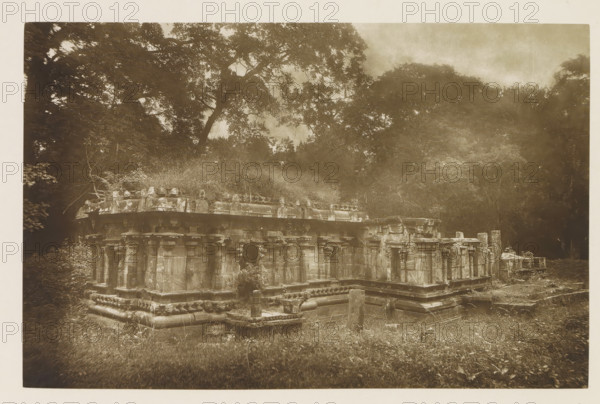 Ruins of the Dalada Maligawa temple at Polonnaruwa, Sri Lanka, page, photomechanical print, title: 'DALADA MALIGAWA AT POLONNARUWA.', photographer: Henry William Cave, Polonnaruwa, cliché maker: anonymous, 1896, South Asia, paper, Woodburygravure, height 124 mm x width 193 mm, Print for page 119., temple, shrine, Hinduism, Buddhism, Jainism, ruin of a building, architecture, A sepia-toned photograph shows a stone structure with ornate columns and carved details, appearing low and rectangular with layered bases and a raised platform; the stonework displays weathered surfaces, rounded moldings, and sculpted elements along the top edge. Surrounding the structure is overgrown grass and low vegetation, with a dense backdrop of tall trees whose branches and foliage form a canopy above and behind the ruins. The scene is framed by darker shaded foliage at the sides, and the sky above the trees is lighter in tone, creating contrast with the silhouetted tree forms.