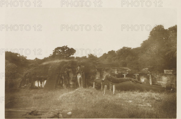 Ruins of the rock temple Gal Vihara at Polonnaruwa, Sri Lanka, page, photomechanical print, title: 'ROCK TEMPLE AT POLONNARUWA.', photographer: Henry William Cave, Polonnaruwa, cliché maker: anonymous, 1896, South Asia, paper, Woodburygravure, height 125 mm x width 194 mm, Print for page 121., temple, shrine, Hinduism, Buddhism, Jainism, sculpture (relief, sculpture), Gal Vihara, A sepia-toned photograph showing a grassy foreground leading to a rocky hillside with carved stone structures; a standing carved figure is visible against the rock face and a large reclining carved figure lies on the right side supported by short stone pillars; trees and dense foliage rise behind the hill, and the sky above is pale and empty.