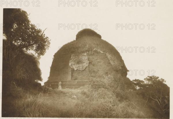 View of the ruins of the Maha Seya stupa, Mihintale, Sri Lanka, page, photomechanical print, The Maha Seya stupa has now been restored., title: 'MAHA SEYA DAGABA.', photographer: Henry William Cave, Ceylon, cliché maker: anonymous, 1896, South Asia, paper, Woodburygravure, height 126 mm x width 195 mm, Print for page 29., temple, shrine, Hinduism, Buddhism, Jainism, ruin of a building, architecture, A large rounded stone structure covered with vegetation dominates the center of the image, with rough masonry visible through patches of grass and plants on its surface and a thicker growth at its top; a narrow dirt path runs along its base and a person in light clothing stands near the structure; tall grasses and shrubs fill the foreground, while trees with dense foliage frame the left edge and appear in the distance at the right, and the sky above is pale and mostly clear.