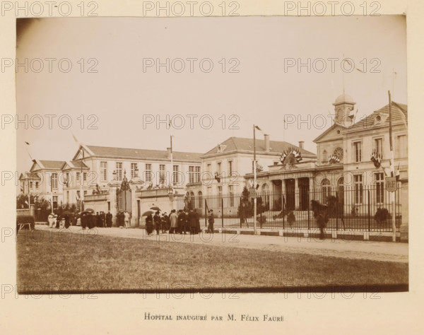 Hôpital inauguré by M. Félix Faure, Facade of the Hôpital Samuel Pozzi in Bergerac, inaugurated by President Félix Faure, photograph, page, Bergerac, 1895-06-04, photographer, paper, daylight gelatin silver print, height 117 mm x width 168 mm, hospital, facade (or house or building), A sepia-toned photograph showing a large classical building complex with multiple connected wings, tiled roofs, decorative pediments, and a central entrance with columns and ornate sculptural details above it, flanked by tall windows and a dome-like cupola on one section; a wrought-iron fence and gate run across the front with flagpoles and flags displayed along the façade; a group of people dressed in early twentieth-century clothing gather and stand near the entrance and along the sidewalk, some holding umbrellas; a grassy foreground fills the lower part of the image and the sky above is mostly clear.