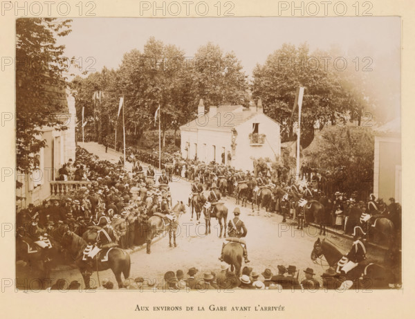 Bergerac (résumé historique) ses hotels illustres, book, publisher: Imprimérie Générale du Sud-Ouest (J. Castanet), Bergerac, 1895, paper, printing, daylight gelatin silver printing, height 252 mm x width 167 mm x thickness 16 mm, A wide street scene shows a crowd of people lining both sides of a road flanked by buildings and tall trees, many individuals wearing hats and period clothing, with mounted soldiers in uniform on horses forming a procession down the center of the street, flags on poles rising above the crowd, a two-story white building with a small balcony and shutters visible on one side, and dense foliage and trees filling the background.