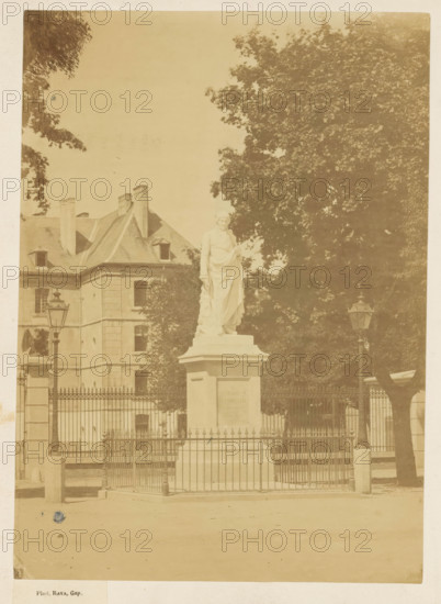 Statue of Charles-François de Ladoucette by Jean Marcellin in Gap, photograph, page, photographer: Rava, Gap, 1866, Provence-Alpes-Côte d'Azur, paper, albumen print, albumendruk, height 219 mm x width 156 mm, Person, Charles-François de Ladoucette, monument, statue, A pale photograph shows a stone statue of a robed figure standing on a rectangular pedestal behind a decorative metal fence, flanked by two tall ornate lampposts. Behind the statue is a large tree with dense foliage and to the left a multi-story building with a sloped roof, chimneys, and small dormer windows. The foreground shows a paved area and the fence includes pointed finials and gate posts. The image has a faded, sepia tone.