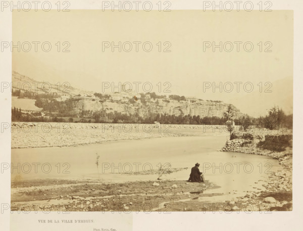 Vue de la Ville d'Embrun, View of Embrun, photograph, page, photographer: Rava, Embrun, c. 1856 - in or before 1866, Rava, photographer, Provence-Alpes-Côte d'Azur, paper, albumen print, height 157 mm x width 219 mm, city-view in general, 'veduta', river, A sepia-toned riverside scene showing a wide calm river with a rocky shore and a low pebbled embankment, a distant elevated town on a cliff with clustered buildings and a prominent tower, tree lines along the cliff base, terraced hills rising beyond, scattered rocks and sparse vegetation in the foreground, and a solitary seated figure wearing a dark coat and hat near the water's edge.