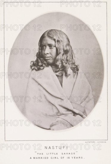 Nastufi 'the little savage' a married girl of 16 years, page, photomechanical print, number: 'No. 25', photographer: Bourne & Shepherd, Nilgiris, photographer: Nicholas & Curths, cliché maker: anonymous, printer (person): Autotype Printing Company, London, c. 1863 - in or before 1873, printer (person), paper, collotype, photoprint, height 122 mm x width 98 mm, adolescent, young woman, maiden, A sepia-toned oval portrait of a young person with long wavy hair wearing layered necklaces and wrapped in a light-colored blanket, shown from the torso up with a neutral expression and eyes looking slightly to the side, framed by a rectangular border on a pale background.