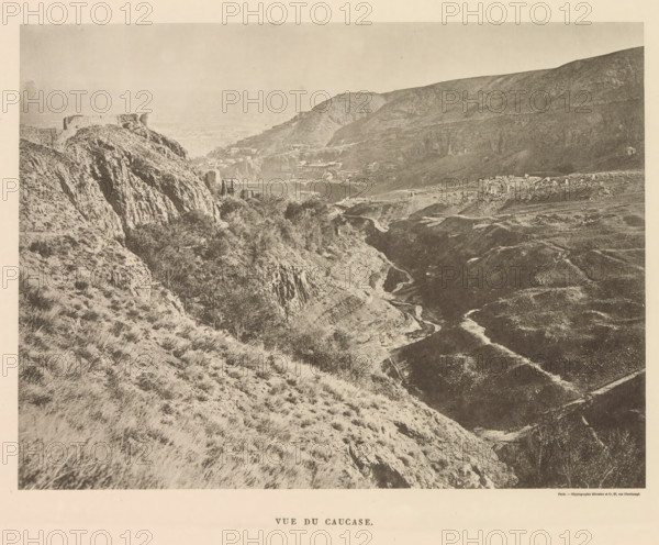 Vue du Caucase, View of the Caucasus, page, photomechanical print, name: 'Silvestre et Cie, 97, rue Oberkampf.', printer (person): Silvestre et Cie, Paris, c. 1869 - in or before 1885, printer (person), paper, collotype, photoprint, height 129 mm x width 174 mm, mountains, Caucasus, A wide landscape photograph showing rugged hills and steep rocky cliffs with sparse grasses and scattered shrubs, a winding dirt path or dry streambed cutting through a valley, clusters of trees and denser vegetation in lower areas, terraces or layered slopes on the distant hills, small buildings and structures nestled along the mid-distance slopes, and a prominent flat-topped rocky outcrop on the left side under a pale sky.
