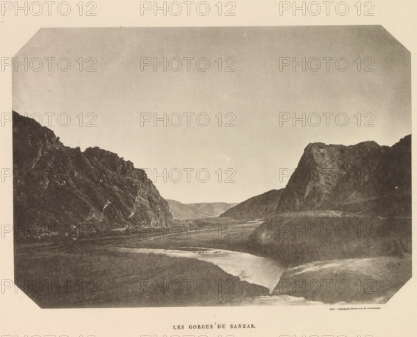 Les Gorges' du Sanzar, View of a gorge in Uzbekistan, page, photomechanical print, name: 'Silvestre et Cie, 97, rue Oberkampf.', printer (person): Silvestre et Cie, Paris, c. 1869 - in or before 1885, Silvestre et Cie, printer (person), paper, collotype, photoprint, height 126 mm x width 179 mm, ravine, chasm, abyss, canyon, A wide landscape photograph showing steep rocky cliffs on both sides with a narrow river winding through the valley between them, a flat plain in the foreground, and an open sky above.