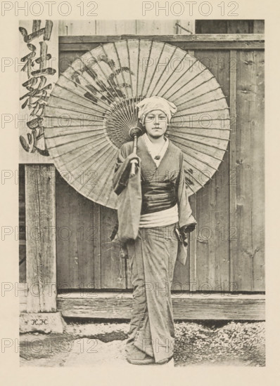 Jeune fille japonaise, Girl with a parasol in Japan, page, photomechanical print, inscription: 'HK', inscription: 'Photographie de l'autoriteit.', photographer: Hugues Krafft, Japan, cliché maker: anonymous, in or after 1882 - in or before 1883, Hugues Krafft, photographer, cliché maker, paper, collotype, photoprint, height 173 mm x width 122 mm, Photo opposite title page., folk costume, regional costume, child, parasol, sunshade, A person standing in front of a wooden wall holding a large paper umbrella behind their head, wearing a headscarf and traditional robe with a wide sash, with visible folds and texture in the fabric and a neutral facial expression, the umbrella showing radial ribs and a darker circular center, wooden posts and planks framing the scene and a gravelly ground beneath the person.
